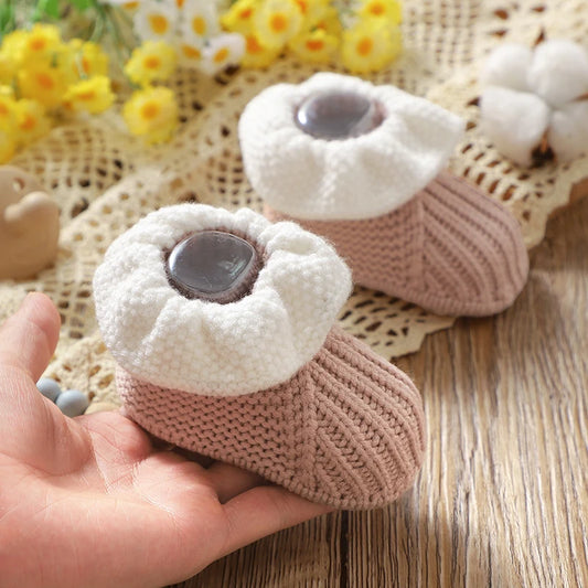 Knitted baby booties with a hand holding one, placed on a textured surface with flowers in the background.