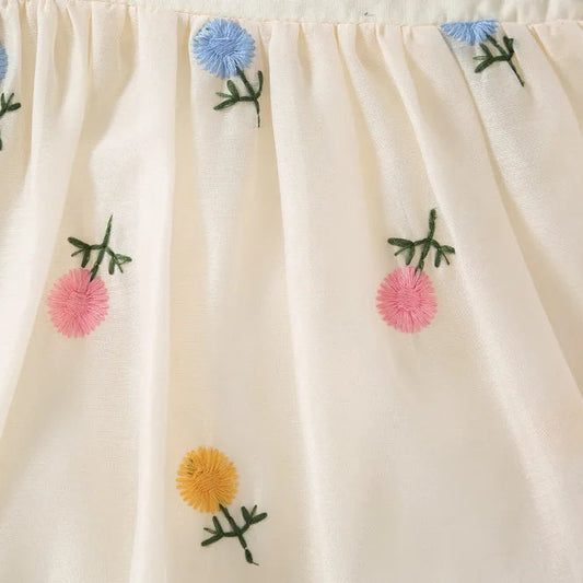 Close-up of artisan floral embroidery on white cotton baby romper featuring light pink, blue, and yellow flowers with green leaf detailing and textured square neckline.