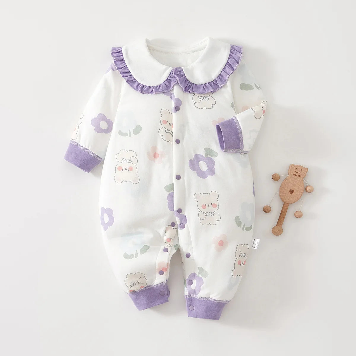 White cotton baby one-piece with a purple teddy bear and flower pattern, featuring a purple Peter Pan collar with ruffle trim and snap button closure.