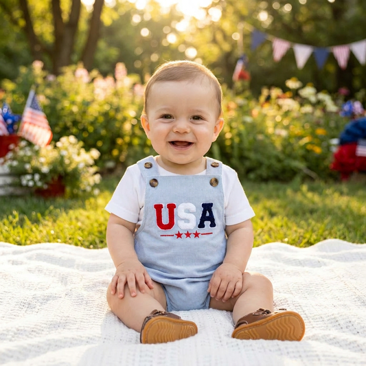 Smiling baby sitting outdoors on a blanket wearing the light blue USA embroidered bubble romper layered over a white short-sleeve t-shirt.
