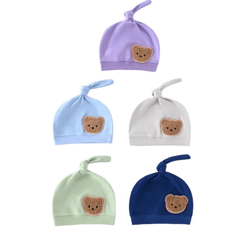 A grouping of five knotted newborn baby hats featuring teddy bear patches in purple, light blue, grey, sage green, and navy blue.