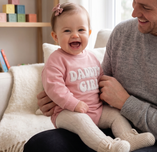 Happy baby girl wearing a pink "Daddy's Girl" long sleeve bubble romper sitting with her dad on a couch.