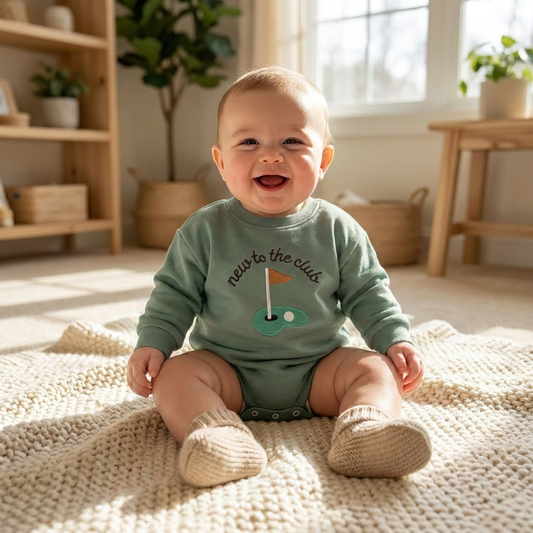 A smiling baby sitting on a textured blanket in a cozy room wearing the green "New to the Club" long-sleeve golf romper.