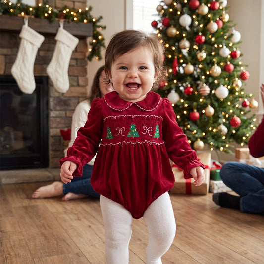 Smiling baby girl wearing a red smocked holiday bubble romper and white tights standing in front of a Christmas tree and fireplace