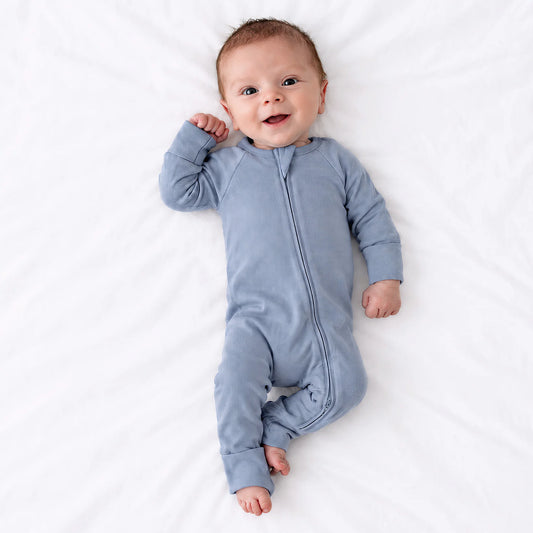 Baby model lying on white sheet wearing Kidsely solid dusty blue bamboo viscose zipper romper sleeper
