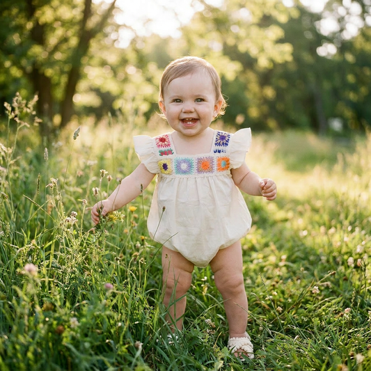 A smiling baby girl wearing the cream-colored crochet flower romper standing in a sunny, green grass field outdoors.