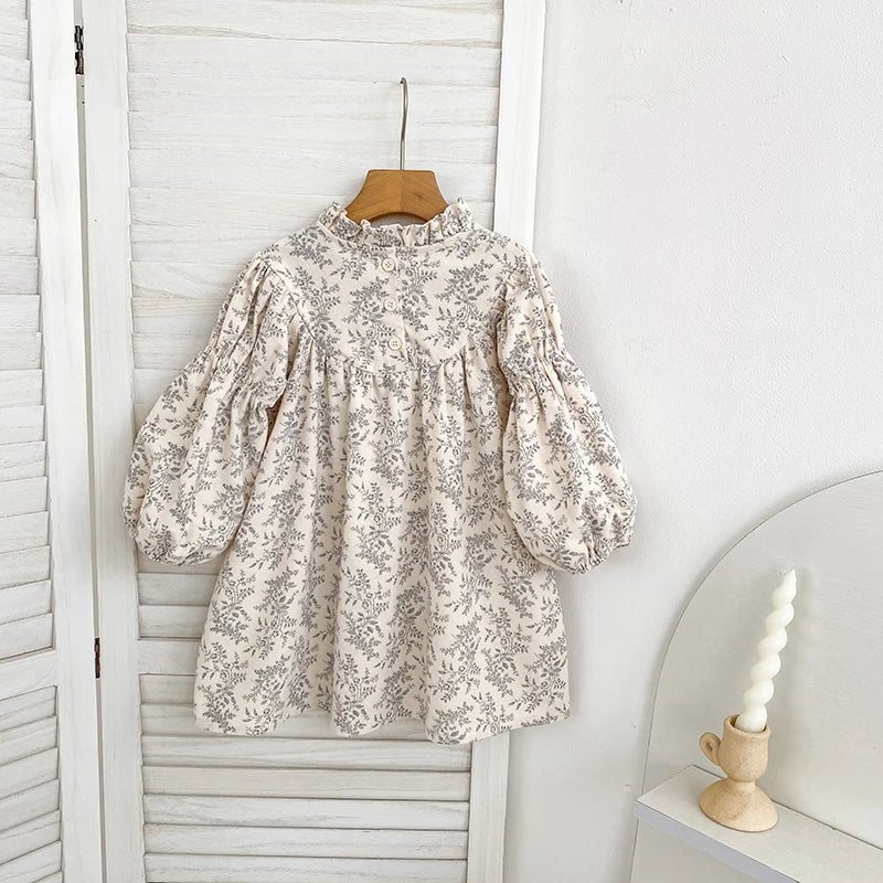The back view of a vintage-inspired girls cream and gray floral dress hanging on a wooden hanger against a white slatted background.