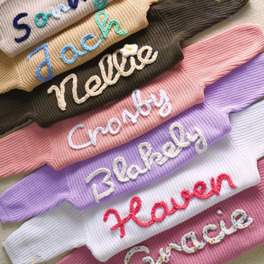 Stack of personalized knit baby sweaters showing custom hand-embroidered names in various font styles and yarn colors