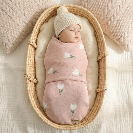 A sleeping newborn baby swaddled safely in a soft pink cotton knit tulip pattern blanket inside a woven Moses basket.