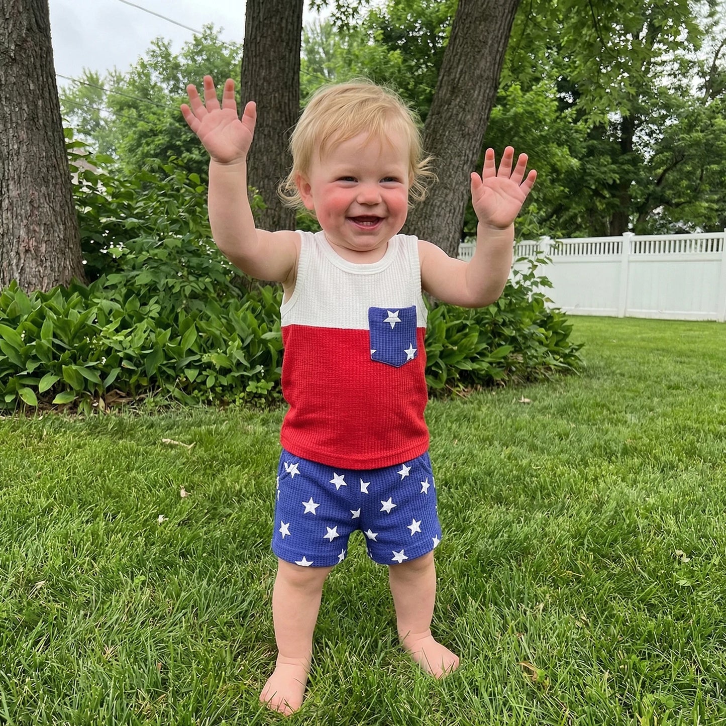 Toddler boy smiling and playing outside in the grass wearing a 2-piece red, white, and blue star-print summer outfit.