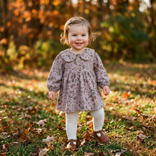 A smiling toddler girl standing in autumn leaves wearing the Kidsely vintage mauve floral dress paired with beige tights.