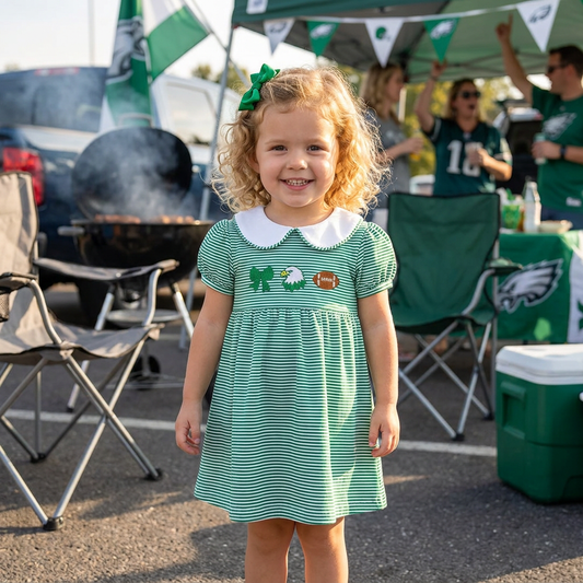 A smiling toddler girl wearing the green striped game day dress standing outdoors at a football tailgate.