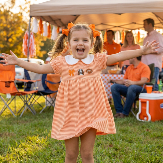 A happy toddler girl with outstretched arms wearing the orange and white striped game day dress with hound dog embroidery and orange hair bows, standing outdoors on the grass at a sunny football tailgate party.