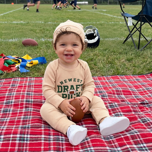 A smiling baby wearing a beige long-sleeve "Newest Draft Pick" football romper and matching knot hat, sitting on a red plaid blanket outdoors holding a small football.
