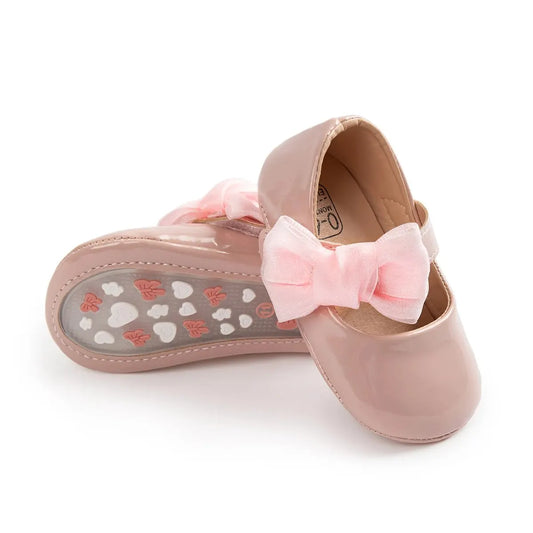 Soft pink patent leather first walker shoes for baby girls featuring a large ribbon bow and butterfly-patterned grip sole.