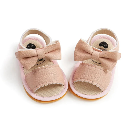 Close-up of pink baby girl first walker sandals featuring a large leather bow knot, scalloped trim, and a flexible tan rubber sole.