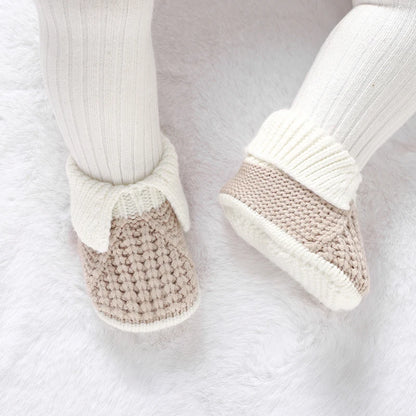 A close-up photograph of a baby's feet wearing beige knitted booties with white ribbed cuffs and white ribbed leggings, resting on a soft white fur surface.