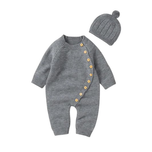 Flat lay of grey knitted baby romper with long sleeves and asymmetrical wooden side buttons, paired with a matching ribbed beanie hat with pom-pom.