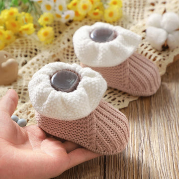 Knitted baby booties with a hand holding one, placed on a textured surface with flowers in the background.