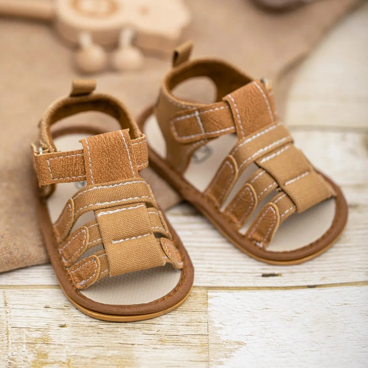 Baby boy first walker sandals in tan brown faux leather with white stitching, featuring a multi-strap fisherman design and non-slip rubber sole for infants and toddlers.