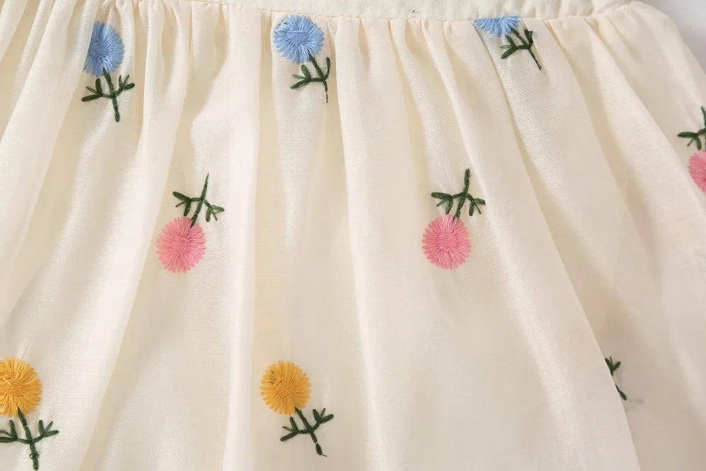 Close-up of artisan floral embroidery on white cotton baby romper featuring light pink, blue, and yellow flowers with green leaf detailing and textured square neckline.