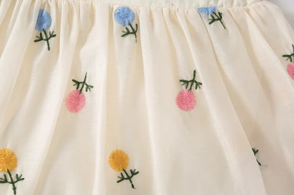 Close-up of artisan floral embroidery on white cotton baby romper featuring light pink, blue, and yellow flowers with green leaf detailing and textured square neckline.