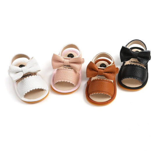 A collection of four pairs of baby girl first walker sandals with decorative bow knots and scalloped edges, shown in white, pink, tan, and black leather with non-slip rubber soles.