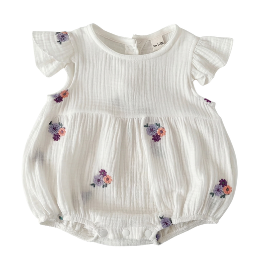 Front view flat lay of a white cotton muslin baby bubble romper featuring purple and peach floral embroidery, ruffle sleeves, and a gathered bodice on a plain background.