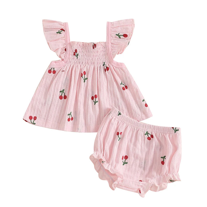 Newborn baby girl summer outfit featuring a blush pink smocked babydoll top with red cherry print, fly sleeves, and matching ruffle trim shorts.