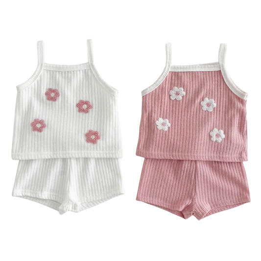 Flat lay of two-piece baby girl summer outfits in white and dusty pink, featuring ribbed knit sleeveless tank tops with 3D floral appliqués and matching shorts.
