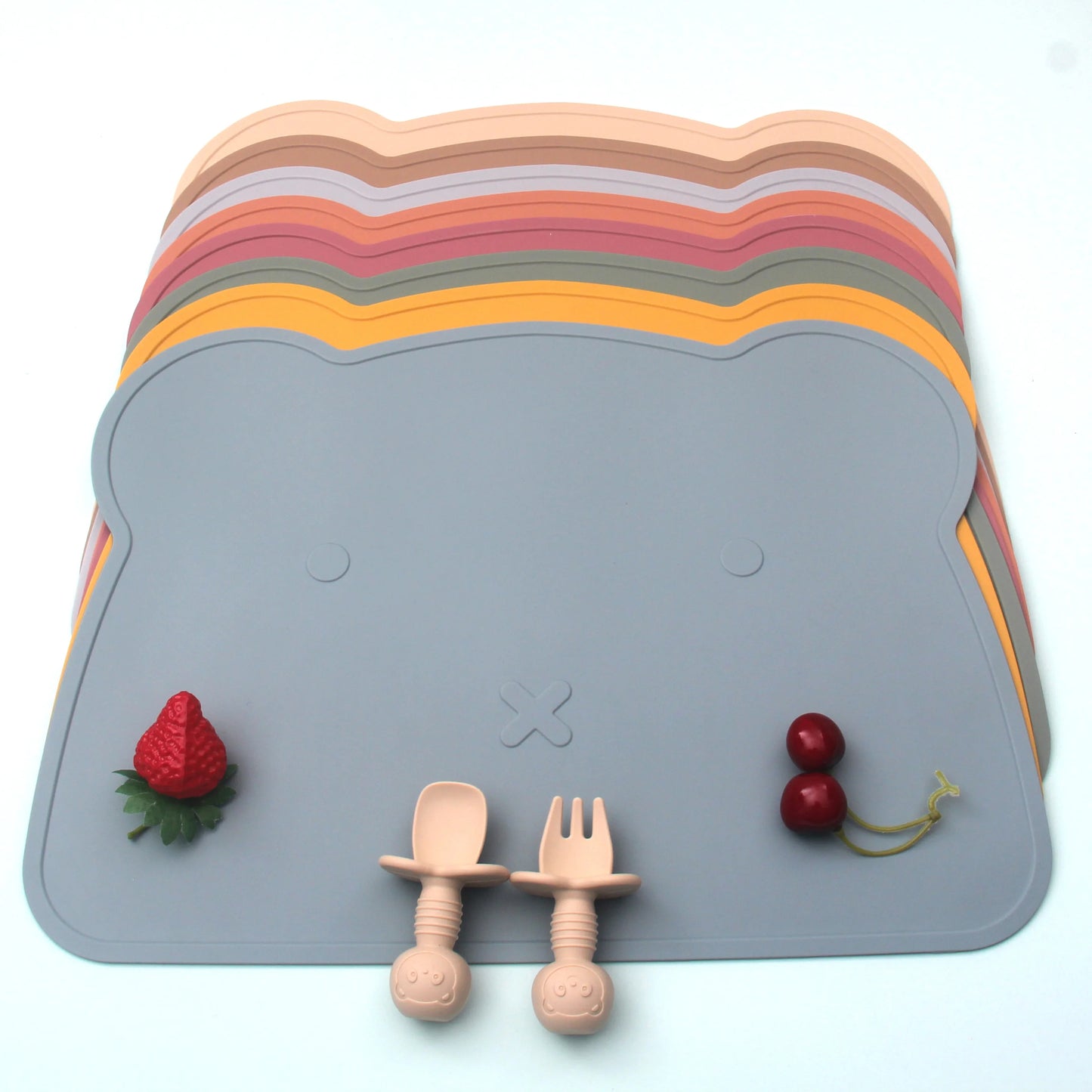 Stack of colorful silicone placemats with a gray base, wooden utensils, and toy fruits on a white background.