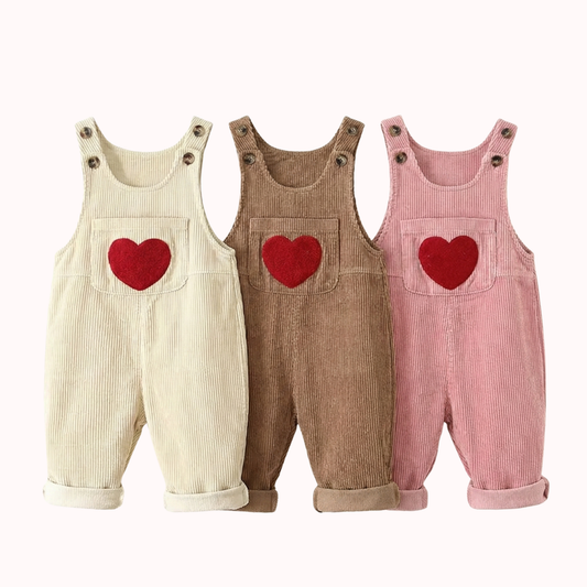 Flat lay product photo of three pairs of toddler corduroy overalls laid side-by-side on a white background. The colors are cream beige, mocha brown, and dusty pink. Each pair features a large, red, fuzzy chenille heart patch on the front chest pocket, two dark buttons on the straps, and cuffed pant legs.