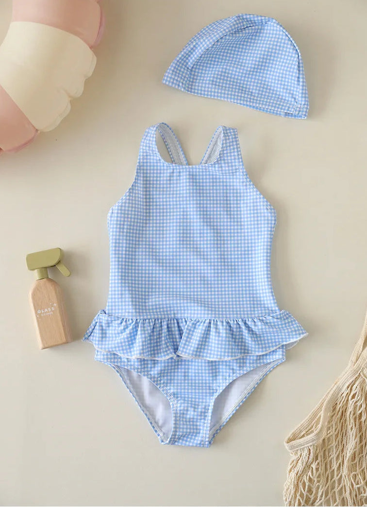 A baby girl's one-piece gingham swimsuit in Cornflower Sky Blue featuring a ruffle skirt and backless design, paired with a matching blue checkered swim bonnet.