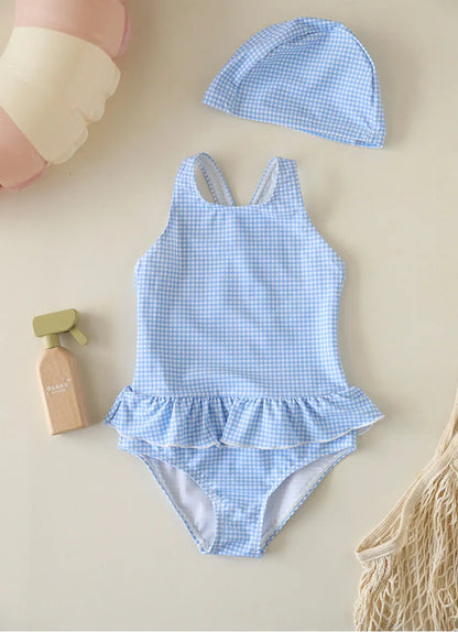 A baby girl's one-piece gingham swimsuit in Cornflower Sky Blue featuring a ruffle skirt and backless design, paired with a matching blue checkered swim bonnet.