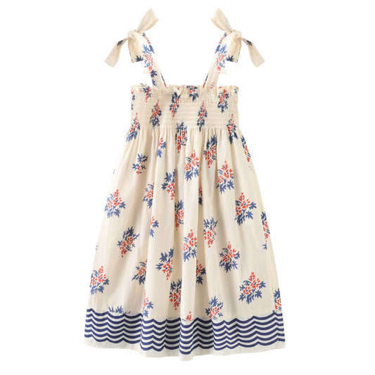Cream-colored 100% cotton girls' sun dress featuring a smocked bodice, adjustable tie-shoulders, and a blue floral print with a wave-patterned hem.