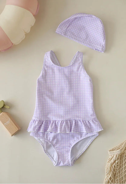 A Wildflower Lavender purple gingham swimsuit set for girls, including a backless one-piece with ruffle trim and a matching purple checkered swim hat.