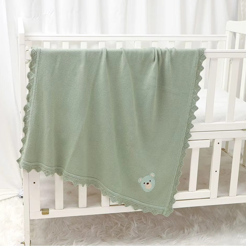 Green baby blanket with a teddy bear design draped over a white crib.