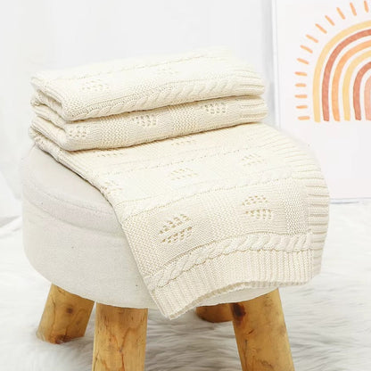 Stack of cream-colored knitted blankets on a wooden stool with a colorful abstract painting in the background.