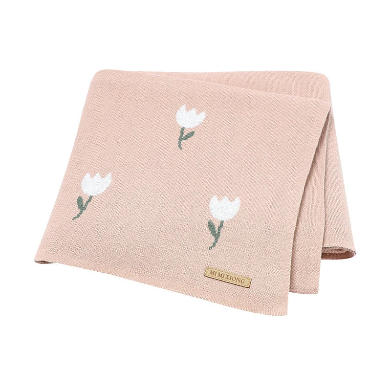 Premium blush pink 100% cotton knit baby blanket featuring a white woven tulip pattern, neatly folded to show the soft texture and brand accent tag.