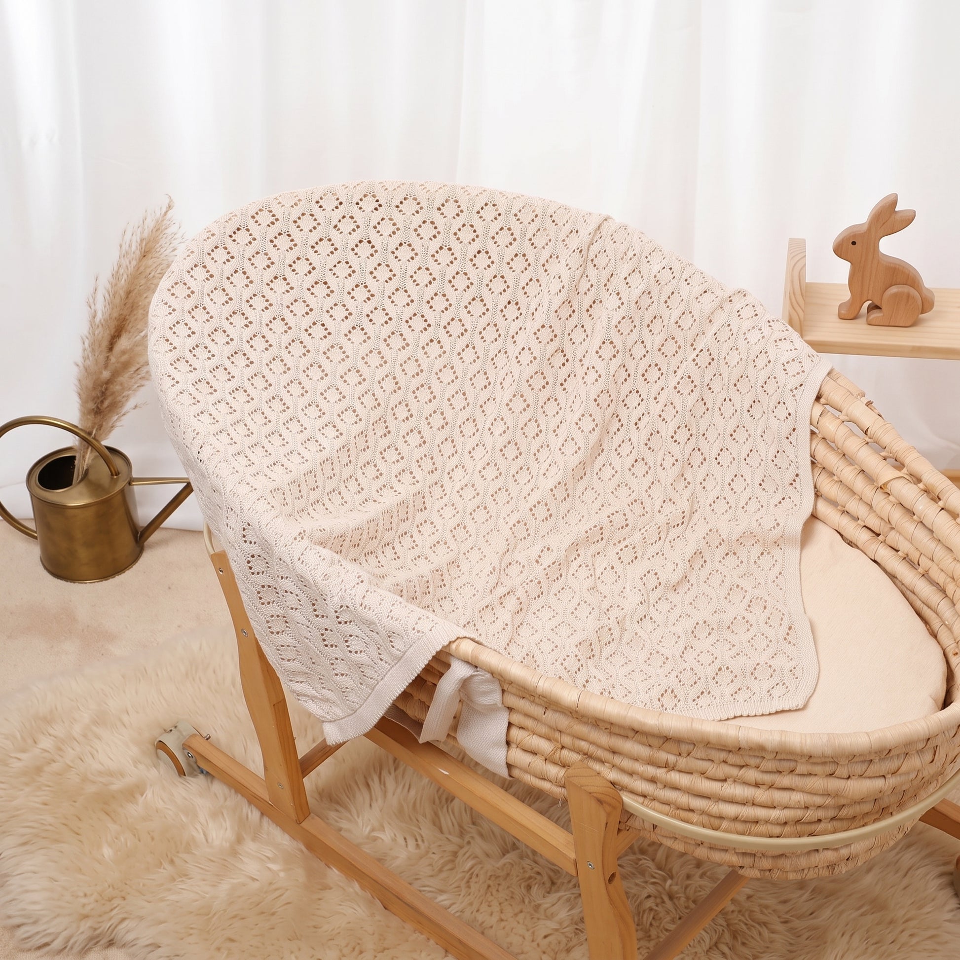 A woven rattan baby bassinet with a oatmeal 100% cotton blanket draped over the sides, styled on a fluffy cream rug with a small wooden rabbit figurine and a brass watering can in the background.