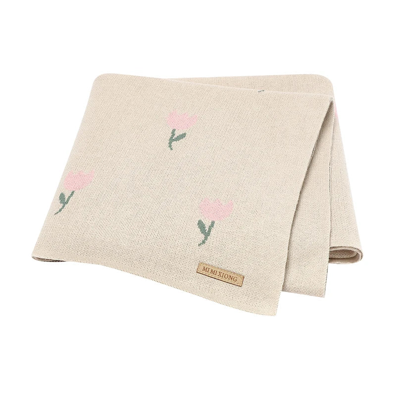 An oatmeal beige neutral cotton knit baby blanket featuring a subtle pink tulip pattern, folded on a clean white background.
