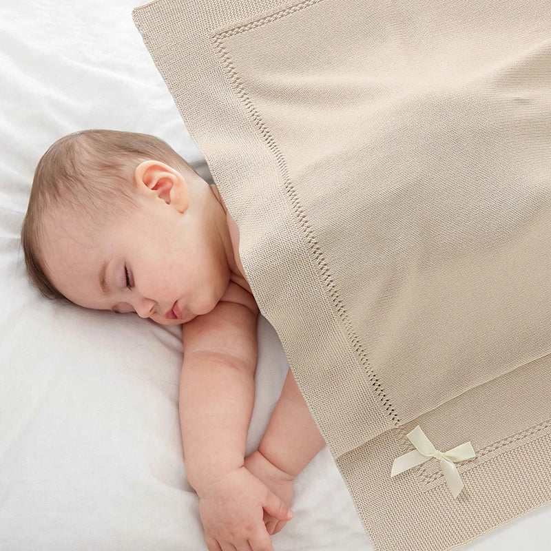Newborn sleeping comfortably under an oatmeal cotton knit baby blanket with bow detail