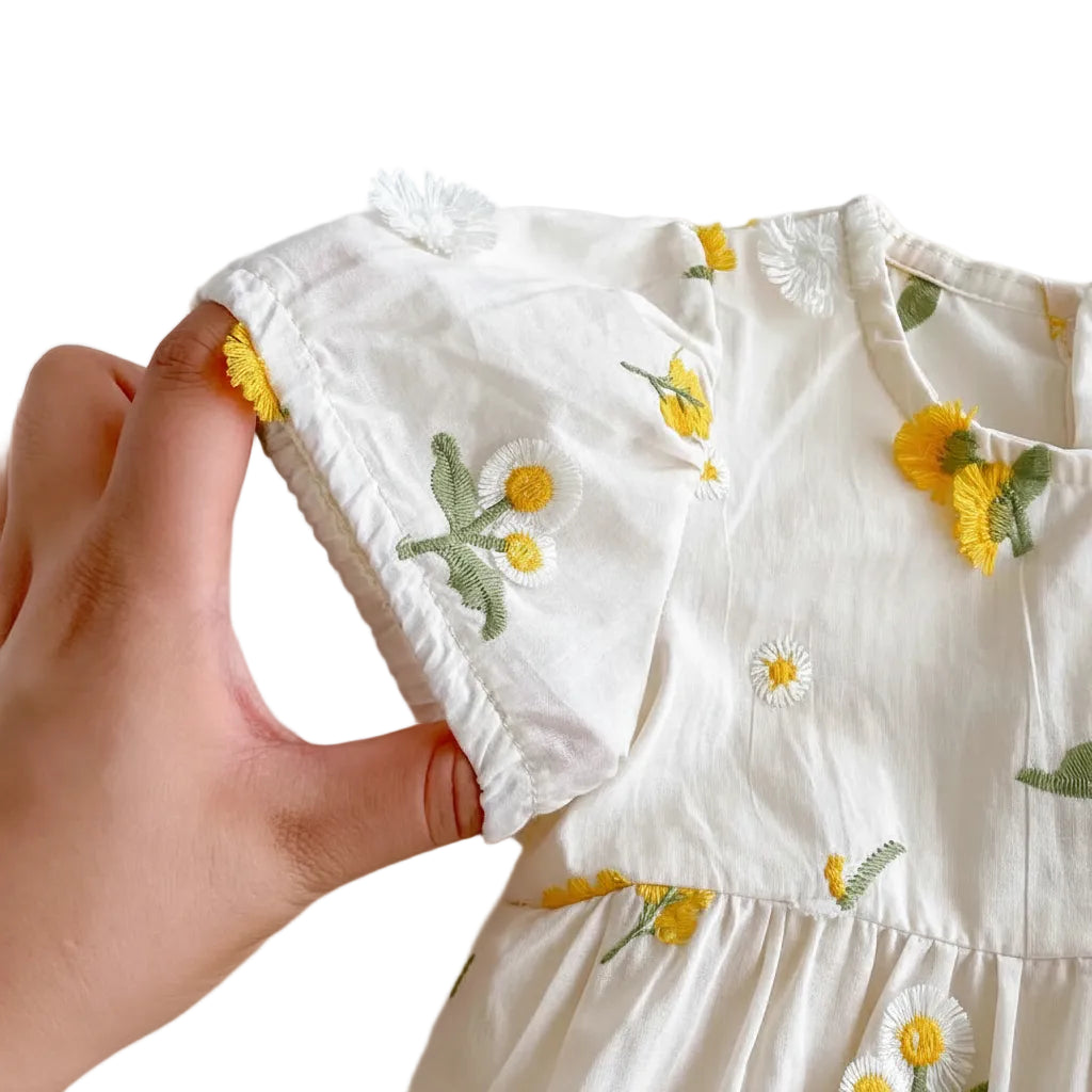 Hand holding the edge of the puff sleeve on a baby girl romper to display the detailed 3D yellow daisy embroidery and soft fabric texture.