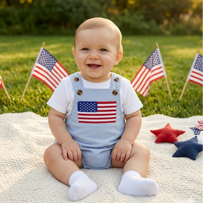 Smiling baby sitting on a picnic blanket outdoors wearing a light blue USA flag patch bubble romper layered over a white t-shirt for a 4th of July outfit.