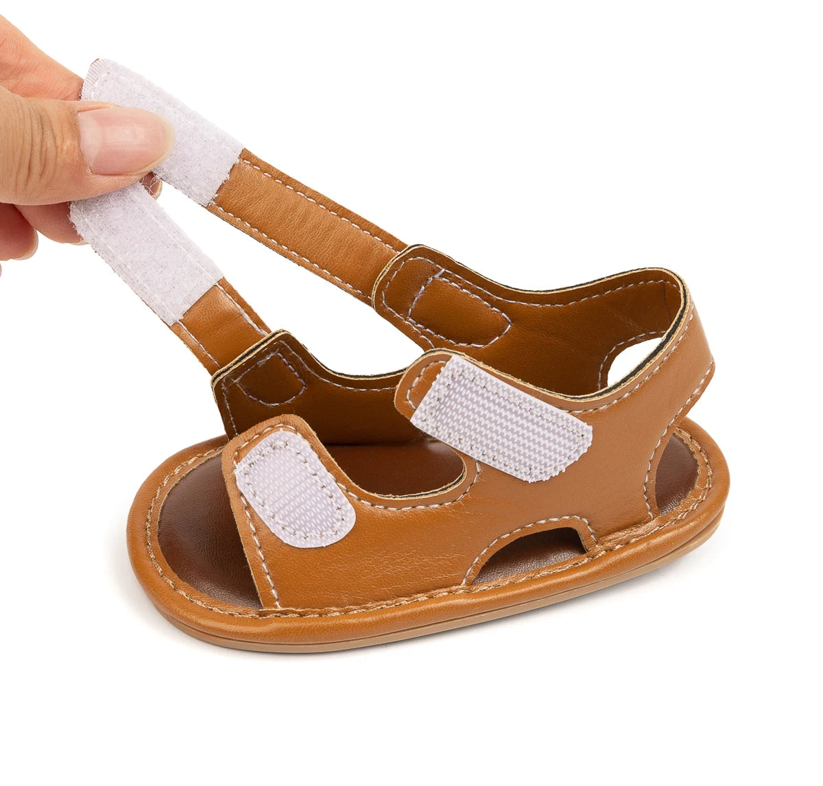 Hand opening the adjustable hook-and-loop strap on a brown baby sandal.