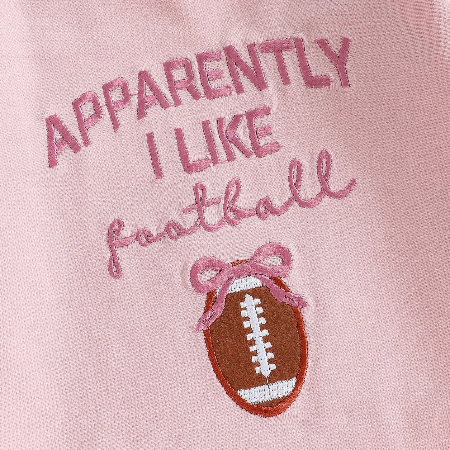 Close-up of the "Apparently I Like Football" pink lettering and the embroidered football graphic with a pink bow detail on the baby romper.