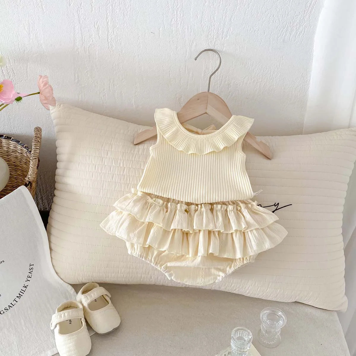The Aria baby girl ruffled set displayed on a wooden hanger against a neutral pillow, showcasing a boutique aesthetic.