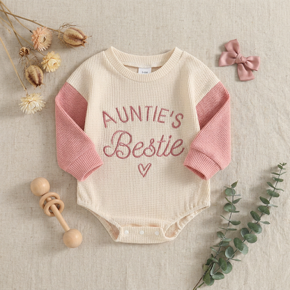 Flat lay of the Auntie's Bestie baby girl bodysuit styled as a gift with a wooden baby rattle, greenery, and a pink hair bow.