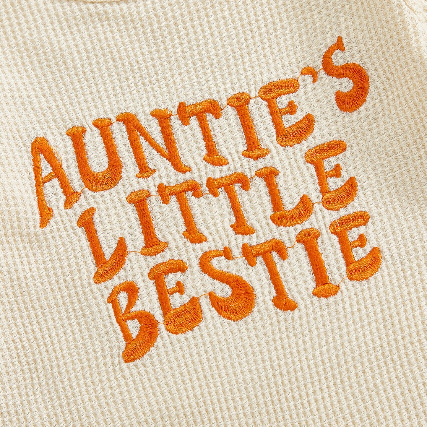 Close-up showing the premium orange textured embroidery of the words "Auntie's Little Bestie" on the beige waffle knit fabric.