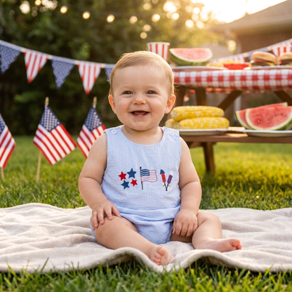 Smiling baby boy wearing the blue striped 4th of July bubble romper sitting on a blanket at an outdoor summer picnic.